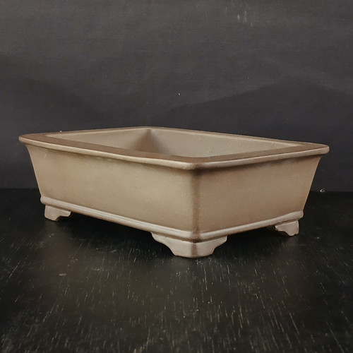 An eye-level studio photograph of a 6-inch unglazed rectangular Yixing ceramic bonsai pot (No. 2482-1). The pot is crafted from traditional Yixing clay, featuring a smooth, matte finish in a warm, earthy taupe or light brown color. Its design is a classic rectangle with clean lines and sides that flare slightly outward toward the top, finished with a subtle, flat rim. The pot stands on four elegant, bracket-style feet at each corner, elevating it off the surface. It is positioned on a dark, textured wooden surface against a solid, dark grey background, with soft lighting from the side that highlights the simple, refined proportions and the natural texture of the unglazed ceramic. This pot's minimalist aesthetic and traditional material make it a versatile choice for various bonsai styles.