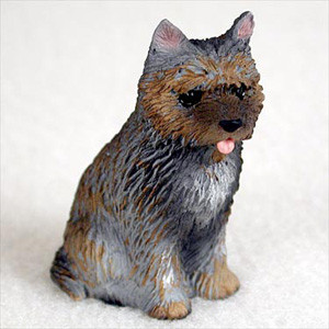 A detailed miniature figurine of a brindle Cairn Terrier, sitting upright on a plain white surface. The small dog features a realistic, textured coat with a blend of dark charcoal gray and warm tan-brown fur, characteristic of the brindle pattern. Its face is primarily brown with a dark muzzle, a tiny black nose, and a small pink tongue sticking out in a playful expression. The figurine has pointed ears and dark, lifelike eyes. Designed as a charming accessory for bonsai trees or miniature gardens, this Cairn Terrier figurine captures the alert and spirited personality of the breed.