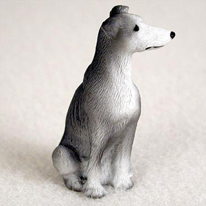 A detailed miniature figurine of a blue Greyhound dog is depicted in a poised sitting position. This small figure is designed as a charming decorative accessory for bonsai tree displays or miniature gardens. The dog features a distinctive "blue" or soft grey coat covering its back and the top of its head, transitioning into a crisp white on its long snout, chest, and front paws. It has the signature slender build of a sighthound, with a long, tapered muzzle, a small black nose, and tiny, dark eyes that give it an alert expression. The surface is subtly textured to represent short, fine fur. Set against a neutral, off-white background, the figurine looks off to the right with its ears tucked back in a relaxed manner, ready to add a touch of life to a miniature landscape.