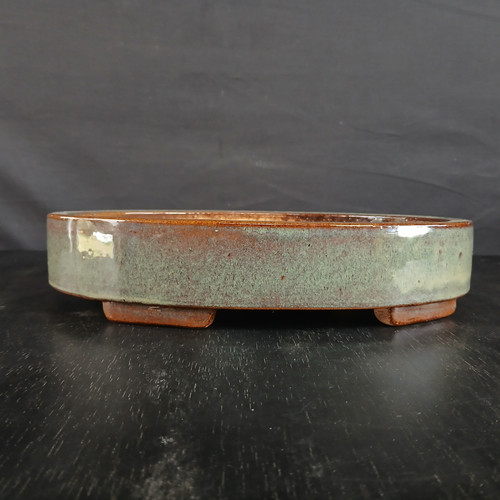 A 12-inch glazed oval Yixing bonsai pot is shown from a low, side-profile angle. The shallow pot features a classic oval shape with straight sides and is elevated on small, blocky feet made of unglazed, reddish-brown clay. 

The exterior of the pot is finished with a stunning, multi-toned glaze. A band of warm, earthy brown clay is visible along the very top rim and at the base. The main body of the pot is covered in a speckled, mottled glaze that blends shades of soft sage green, pale seafoam, and subtle blue-grey. Small darker specks are scattered throughout the glaze, giving it a natural, stone-like texture. The high-gloss finish creates a bright white reflection on its smooth, curved surface on the left. The bonsai pot sits on a dark, textured black wooden surface against a deep charcoal grey backdrop, highlighting the pot's unique colors and craftsmanship.