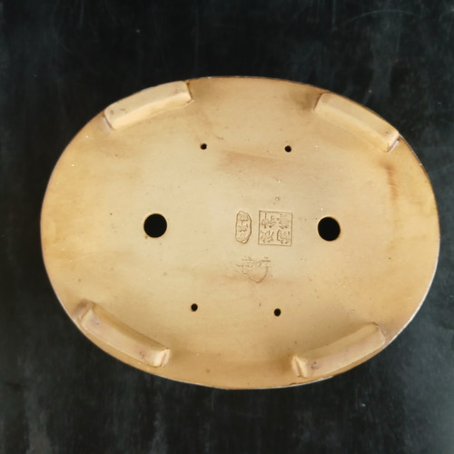 An overhead, close-up view shows the underside of a 12-inch oval Yixing bonsai pot, highlighting its construction and functional details. The unglazed ceramic bottom is a light tan or beige color, featuring two large, circular drainage holes in the center for proper water flow. Surrounding these are four smaller, precisely placed holes used for securing the tree to the pot with bonsai training wire. The pot is elevated by four short, curved rectangular feet positioned near the outer edges. In the center of the pot's base, several authentic Chinese seal marks are stamped into the clay, signifying its origin and craftsmanship from the Yixing region. The pot is set against a dark, textured background that contrasts with the light clay.