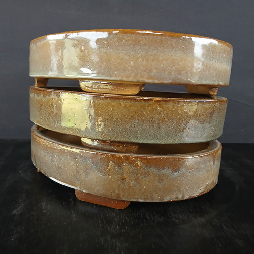 A stack of three 12-inch glazed oval Yixing bonsai pots is presented against a dark, minimalist background. The pots are stacked vertically, showcasing their uniform, shallow elliptical shape and low profile. Each pot features a rich, high-gloss glaze with a mottled, earthy color palette of amber, tan, and dark brown, with subtle hints of olive green and a speckled "oil spot" texture.

The glaze is reflective, catching the light and highlighting the smooth, curved edges of the ceramic. Small, integrated rectangular feet are visible at the base of each pot, designed to provide elevation for proper drainage and airflow. The stack sits on a dark, slightly reflective surface, which emphasizes the warm tones and artisanal quality of the Yixing clay. The overall composition highlights the natural variation in the glazing process, making each pot appear unique while maintaining a cohesive aesthetic.