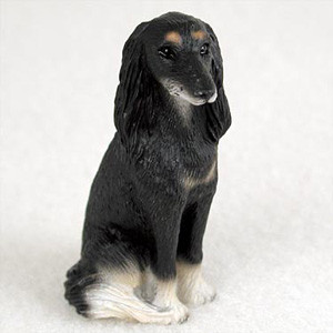 A detailed miniature figurine of a Saluki dog, designed as a decorative accessory for a bonsai tree landscape. The Saluki is shown in a graceful sitting position, looking calmly off to the side. It features a primarily black coat with characteristic tan "eyebrow" spots above its dark eyes and cream-colored fur on its muzzle, chest, and paws. Its long, feathered ears hang elegantly on either side of its slender face, capturing the distinctive and athletic appearance of the breed. The figurine is set against a plain white background, highlighting its fine, hand-painted details and realistic texture.