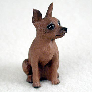 A small, detailed figurine of a red-brown Miniature Pinscher dog, designed as a decorative accessory for a bonsai tree or terrarium. The figurine is captured in a sitting position, looking alertly toward the upper right with its large, pointed ears standing upright. Its dark, glossy eyes and small black nose give it an expressive and lifelike face. The surface of the reddish-brown resin is textured to mimic the short fur of a Min Pin. The dog is posed on a soft, off-white surface that provides a neutral background for the miniature sculpture.