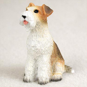 A detailed miniature figurine of a Wire Fox Terrier dog, designed as a decorative accent for bonsai trees or fairy gardens. The small dog is depicted in a sitting pose, looking alertly upward and slightly to the side with a cheerful expression and a tiny pink tongue showing. Its coat is sculpted with realistic wiry fur textures, featuring a predominantly white body, chest, and bearded muzzle, contrasted by warm reddish-tan and brown patches on its head, folded-over ears, and across its back and tail. The figurine sits on a neutral, off-white surface, highlighting its intricate painted details from its dark, inquisitive eyes to the grey-shaded tips of its paws.