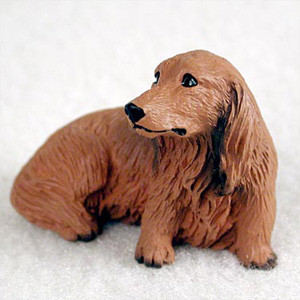 A small, detailed miniature figurine of a long-haired red dachshund, ideal as a decorative accessory for bonsai tree arrangements or miniature gardens. The dog is sculpted in a resting pose, lying on its belly with its head turned thoughtfully to the side. The figurine features a warm, reddish-brown color and intricate texture that realistically mimics the wavy fur of a long-haired breed, particularly on its long, floppy ears and body. It has a long snout with a black nose and small, dark, expressive eyes. The piece is set against a plain, soft white textured background, emphasizing its design as a charming accent piece for plant enthusiasts.