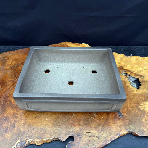 A high-angle studio photograph features a twelve-inch rectangular Yixing bonsai pot, highlighting its professional-grade construction and unglazed, earth-toned ceramic finish. The pot, known for its high-quality purple clay or zisha material, displays a matte, slightly grainy texture in a deep brown-gray hue. Its design includes a subtle recessed rectangular panel on the front face, adding a touch of classic elegance.

Inside the pot, several large drainage holes and smaller holes for anchoring wire are visible, demonstrating its practical use for bonsai cultivation. The rectangular pot is displayed on a large, polished wood slab with a natural, live edge and intricate, swirling grain patterns. This setting is placed against a stark, dark background, which emphasizes the clean lines and sturdy form of the Yixing clay container. This image captures the essential aesthetic and functional qualities of a premium bonsai pot used by enthusiasts.