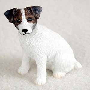 A detailed miniature figurine of a Jack Russell Terrier with a white and brown rough coat, designed as a decorative accessory for a bonsai tree. The small, realistic dog is captured in a sitting pose, featuring a textured, wire-haired appearance that mimics the breed's natural coat. Its head is adorned with dark brown and black patches around the eyes and folded-over ears, while the body remains pure white. The figurine is photographed on a light-colored, sandy surface, showcasing how it can be used to add a touch of life and character to miniature landscapes and indoor bonsai displays.