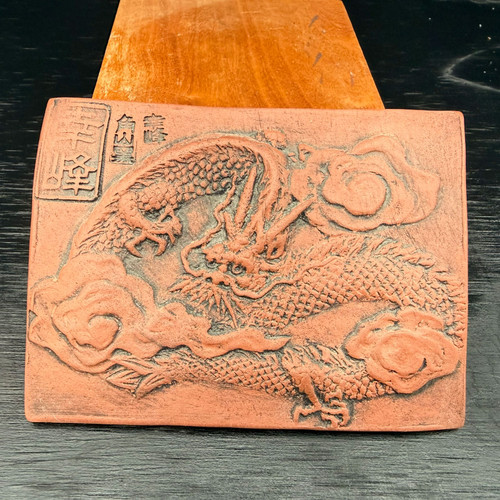 An intricate, rectangular Tokoname clay dragon plaque, hand-carved in high relief by artist Kouji Watanabe. The unglazed red clay features a dynamic traditional dragon weaving through stylized, swirling clouds. The dragon is highly detailed, showing individual scales along its body, sharp claws, and flowing whiskers. In the upper left corner, several Japanese characters are embossed, including a set within a rectangular border seal. The 4.25-inch ceramic piece has an earthy, matte texture and is shown propped up by a wooden wedge against a dark, wood-grained background. The lighting emphasizes the depth of the relief carving, casting shadows that bring the mythical creature to life.