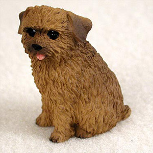 A detailed, miniature Norfolk Terrier bonsai tree figurine, meticulously crafted to serve as a charming accessory for indoor gardens. The small brown dog is depicted in a seated position, looking forward with a playful expression. Its body features a deeply textured, scruffy coat in shades of earthy brown, realistically mimicking the wiry fur of a true Norfolk Terrier. The figurine has small, dark, glistening eyes, a tiny black nose, and a small pink tongue peeking out from its mouth. Its signature V-shaped ears are folded neatly forward. This durable resin figurine is designed to add a touch of personality and life when placed at the base of a bonsai tree, within a succulent arrangement, or as part of a miniature fairy garden display. The dog sits on a plain, light-colored background that emphasizes its intricate, handcrafted details.