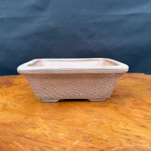 An eye-level, side-profile photograph of a 6-inch rectangular bonsai pot handcrafted by artist Reiho (Katsushi Kataoka) at the Seizan Kiln. The unglazed ceramic pot is an earthy, reddish-tan color and features a unique, intricate texture on its exterior walls that resembles crackled clay or a delicate scale-like pattern. It has a wide, flared top rim with a rough, granular finish and tapers down to a smaller base supported by four small, integrated feet. The pot is positioned on a polished, warm-toned wooden surface against a dark, out-of-focus background, highlighting its artisanal details and rugged, natural aesthetic.