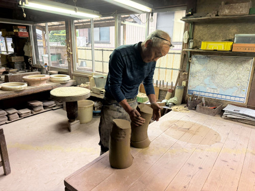 An elderly master potter, likely Katsushi Kataoka (known professionally as Reiho), works inside the Seizan Kiln studio. He is leaning over a large, light-colored wooden workbench, his hands covered in earth-toned clay as he meticulously positions two thick, curved slabs of moist brown clay. The potter has short white hair with glasses resting on his forehead and wears a dark, long-sleeved textured shirt and a heavily soiled, light-colored apron. 

The rustic workshop is filled with natural light from large windows in the background. To the left, several round, unglazed pottery basins in various stages of completion sit on low shelves and a pottery wheel table. To the right, the wall features a map of the Tokai region of Japan, alongside shelves holding tools, a yellow storage bin, and a collection of brushes. The scene captures a moment of focused craftsmanship, illustrating the traditional manual process used to create high-quality bonsai pots.