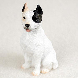 A detailed miniature figurine of a Pit Bull Terrier, designed as a decorative accent for a bonsai tree. The small dog figurine is depicted in a sitting position, facing forward with a gentle expression. Its body is primarily white with a finely textured finish suggesting short fur. A prominent, large black patch covers its left eye and its entire left ear, while the right ear is a contrasting light tan or peach color. The dog has a small black nose and its mouth is slightly open, revealing a tiny pink tongue. This realistic Pit Bull Terrier figurine sits on a plain, light-colored background, showcasing its craftsmanship as a charming addition to a miniature garden or indoor bonsai display.