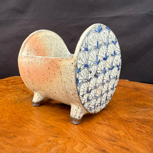 A 4.25-inch Seizan Kiln sculptural accent pot, designed for kusamono or companion planting, sits on a polished wooden table. The ceramic vessel has a unique, elongated cradle shape with two high, semi-circular ends and is supported by four small, stout feet. 

One of the circular end faces is decorated with an intricate, hand-painted blue and white pattern of overlapping arcs, reminiscent of traditional Japanese wave or fish-scale motifs. The rest of the pot’s surface is finished in a speckled, cream-colored glaze with soft orange-tan blushes and tiny dark spots, providing a natural, rustic texture. The pot is displayed on a rich, warm-toned wood surface with a prominent grain against a solid black background, which accentuates its artisanal details and sculptural form.