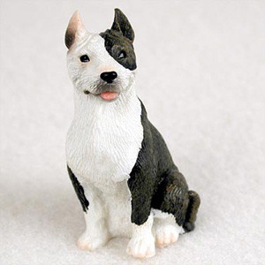 A detailed miniature figurine of a brindle and white Pit Bull Terrier, designed as a decorative bonsai tree accessory. The small dog statue is in a sitting pose, featuring a split-color face with one side dark brindle and the other white. It has alert, upright ears and a friendly expression with its pink tongue slightly visible. The figurine's textured surface realistically depicts short fur, making it a charming addition to a bonsai garden or indoor planter.