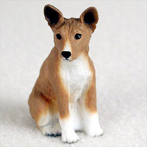 A detailed miniature figurine of a Basenji dog, designed specifically as an accessory for bonsai tree displays, terrariums, or indoor gardens. The small dog is depicted in a seated, alert position, looking slightly to the side with its characteristic large, upright ears. The figurine is hand-painted in a realistic tan and white color scheme, featuring a bright white chest, four white paws, and a thin white blaze on its face. It has dark, expressive eyes and a small black nose, with subtle molded textures across the body to represent short fur. This collectible Basenji miniature is shown against a soft, neutral-colored background, emphasizing its small scale and fine craftsmanship for pet lovers and bonsai enthusiasts alike.