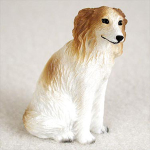 A small, detailed resin figurine of a Borzoi dog, designed as a decorative accessory for a bonsai tree or miniature garden. The dog is depicted in a sitting position, facing toward the right. It features the breed's characteristic slender snout, long, floppy ears, and an elegant, lean frame. 

The figurine is primarily white with warm, golden-tan patches covering its ears and the top of its head, as well as a light tan wash along its back and hindquarters. The surface is intricately textured with fine, sculpted lines to represent the look of long, wavy fur. Small, hand-painted details include black beads for the eyes, a black nose, a thin black line for the mouth suggesting a slight smile, and dark grey accents on the tips of its paws to represent claws. The figurine sits on a soft, off-white textured surface against a neutral, blurred background, highlighting its delicate craftsmanship.