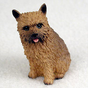 A detailed miniature figurine of a Norwich Terrier, captured in a sitting pose and looking slightly upwards. The small dog statue features a realistic, textured, reddish-brown wiry coat characteristic of the breed, with darker shading around its muzzle. It has alert, pointed ears, large dark expressive eyes, and its tiny pink tongue is peeking out from its mouth in a playful expression. This charming bonsai tree figurine is designed as a decorative accessory for miniature landscapes, fairy gardens, or succulent pots, serving as a perfect collectible for Norwich Terrier enthusiasts.