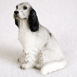 A detailed miniature bonsai tree figurine of an English Setter in the classic Blue Belton color pattern. The small, realistic dog is depicted in a sitting position, looking curiously upward and slightly to the left. Its coat is primarily white with distinctive dark grey ticking and flecks, known as "belton" markings, across its body and paws. The figurine features long, floppy black ears with a wavy, fur-like texture and dark grey patches around its eyes and muzzle. Designed as a charming decorative accent for bonsai displays, the figurine is crafted with intricate details to mimic a dog's fur and is shown against a neutral, soft white background.