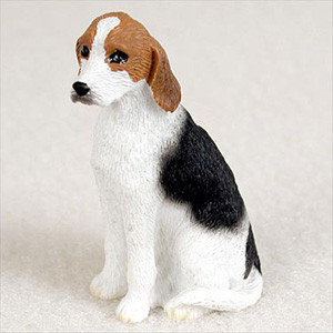 A detailed miniature figurine of an American Foxhound, designed as a decorative accessory for a bonsai tree arrangement. The small dog is captured in a classic sitting pose, looking alertly toward the left. It features a traditional tricolor coat with a crisp white base on its muzzle, chest, legs, and belly. Warm tan markings cover its long, drooping ears and the top of its head around its dark eyes, while a large, deep black patch extends across its back and upper hindquarters. The figurine's surface is finely textured to mimic the appearance of short fur. This tiny collectible is shown against a neutral, off-white background, highlighting its realistic features and suitability for adding scale and character to a miniature landscape or bonsai display.