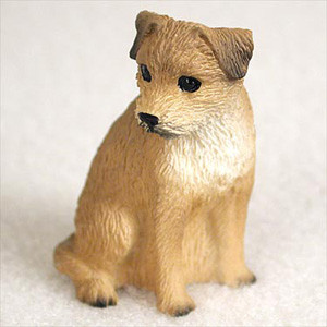 A highly detailed, miniature figurine of a Border Terrier dog, specifically designed as a decorative accessory for a bonsai tree or miniature garden. The figurine depicts the terrier in a calm, sitting pose, looking off to its right. Its coat is a mix of tan and light brown tones, with a lighter cream-colored patch on its chest, and darker brown, folded V-shaped ears. The surface of the figurine is meticulously sculpted to mimic the characteristic wiry and rough texture of a Border Terrier's fur. It features small, dark, expressive eyes and a black nose. The figurine is shown on a clean white surface, highlighting its intricate craftsmanship and realistic appearance.