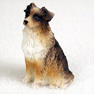 A detailed miniature figurine of a brown and white Australian Shepherd dog, designed as a decorative accessory for a bonsai tree arrangement. The realistic figurine depicts the dog in a classic seated pose, looking attentively off to the side. It features a tricolor coat with a fluffy white ruff around the neck and chest, tan coloring on the muzzle and legs, and dark brown to black patches around the eyes and on the folded ears. The sculpting showcases an intricate, wavy texture that mimics the thick, double coat of an actual Australian Shepherd. The figurine is set against a simple, textured off-white background, emphasizing its lifelike details and hand-painted finish.