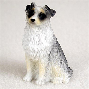 A detailed miniature figurine of a blue merle Australian Shepherd dog, crafted as a bonsai tree accessory. The figurine is depicted in a sitting position, facing slightly to its left. Its coat is intricately textured to mimic fur, painted with a base of crisp white and adorned with mottled grey and black patches characteristic of the blue merle breed. Dark black markings encircle its eyes, giving it a mask-like appearance, complemented by a small black nose and gentle tan accents on its muzzle and feet. This small, lifelike statue is designed to bring personality and a sense of scale to bonsai displays and miniature indoor gardens. It is shown against a simple, off-white backdrop that emphasizes its fine details.
