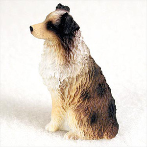 A detailed miniature figurine of an Australian Shepherd dog, captured in a sitting profile view facing left. The figurine showcases the breed's classic tri-color coat, featuring a light tan snout and legs, a thick white ruff around the neck, and a body mottled with shades of brown, tan, and dark black in a merle pattern. The surface is intricately molded to mimic the appearance of long, wavy fur. The dog has a black nose and alert, slightly folded ears. Consistent with the breed standard, it has a docked tail. This decorative bonsai tree accessory is displayed against a clean, off-white background.