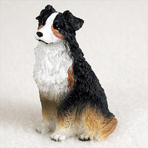 A detailed bonsai tree figurine of a tricolor Australian Shepherd dog, shown sitting upright on a white, textured surface that resembles fine gravel or sand. The small, handcrafted statue captures the breed's distinct characteristics with a black, white, and tan coat. The dog has a prominent white blaze on its forehead and muzzle, small copper-colored markings above its eyes and on its cheeks, and a thick, wavy white ruff around its neck and chest. Its ears and back are primarily black, transitioning into a warm tan or copper color on its lower legs and hindquarters. The figurine is intricately sculpted with a textured finish to mimic the appearance of long, thick fur, and it is positioned in a three-quarter view looking slightly to the left with an alert and loyal expression.