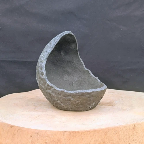 A handcrafted, dark slate-gray concrete planter with a unique, organic shape is positioned on a light-colored wood slab against a dark, textured background.

The planter resembles a broken eggshell or a hollowed-out geode. It features an asymmetrical design where one side rises into a tall, curved peak, while the opposite edge curves down into a lower rim. This creates a deep, protected interior space.

The exterior surface is rugged and craggy, with a coarse texture filled with pits and ridges that mimic the appearance of natural weathered rock or volcanic stone. In contrast, the interior is smoother but retains a fine, porous quality. The lighting highlights the planter's varied textures, casting soft shadows and bringing out light gray highlights along its irregular edges.

The planter sits in the center of a circular, light-toned wooden base with a natural grain. The background is a solid, dark charcoal-colored fabric, which provides a stark contrast that makes the sculptural form of the object stand out.