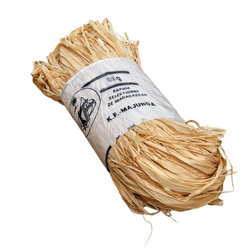 An overhead studio shot of a 50-gram bundle of natural Madagascar raffia, a traditional material used as a bonsai tree branch protection and wiring aid. The bundle consists of numerous long, flat, straw-like strands of dried palm fiber in a natural light tan or golden beige color. A white paper label is wrapped tightly around the center of the bundle to keep the strands organized. The label features black text, including "50g" at the top, followed by "RAPHIA SELECTIONNE DE MADAGASCAR" and "K.F.-MAJUNGA" below. To the left of the text is a small circular logo depicting a tropical scene with a palm tree. The fibrous, flexible texture of the raffia is visible, with loose ends protruding from the sides of the bundle, illustrating its suitability for soaking and wrapping around delicate bonsai branches before applying heavy shaping wire to prevent the bark from splitting.