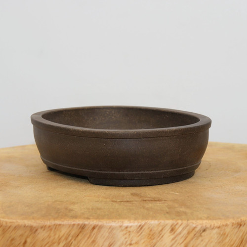 A professional-grade oval unglazed ceramic bonsai pot in a deep, earthy dark brown. This classic bonsai container is shown centered on a rustic, light-grained natural wood slice against a clean, off-white background. The pot features smooth, curved sides with a subtle horizontal line detailing near the rim and integrated drainage feet at the base to ensure optimal root health for a bonsai tree. The matte finish and simple design make it a versatile choice for a variety of bonsai species.