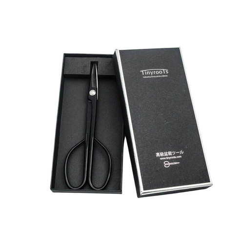 A pair of professional-grade Tinyroots carbon steel long-handled bonsai shears, also known as Hasami shears, are displayed in an open, black foam-lined presentation box. The shears feature a matte black finish with long, slender handles that end in large finger loops, and short, sharp blades joined by a silver pivot screw for precision. 

Leaning against the right side of the box is its black lid, which displays white branding. The lid features the "Tinyroots" logo at the top, the tagline "Cultivating strong root for a lifetime," and at the bottom, the Japanese text "高級盆栽ツール" (High-grade bonsai tool), the website URL "www.tinyroots.com," and a small white icon of the shears. These specialized shears are designed for delicate bonsai tasks such as defoliating and cutting small branches with ease.