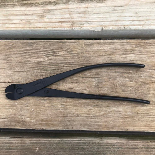 A top-down view of a pair of Tinyroots traditional bonsai wire cutters crafted from forged carbon steel. The professional-grade tool is featured in a sleek matte black finish with long, slender handles that provide leverage and precision. The specialized cutting head has a short, rounded-nose design, specifically engineered to snip aluminum or copper training wires close to a tree's trunk or branch without damaging the delicate bark. The tool rests horizontally across light-toned, weathered wooden planks with a prominent wood grain, highlighting the "Tinyroots" brand name embossed near the pivot point.