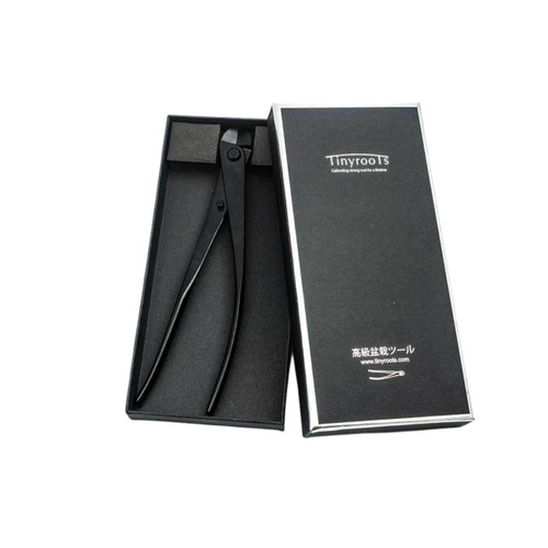 A high-quality pair of Tinyroots carbon steel traditional bonsai wire cutters is presented in a sleek black gift box against a plain white background. The wire cutters feature long, slender black handles and a compact, rounded cutting head, specialized for cutting training wire close to a bonsai tree's branch without causing damage. To the right, the matching black box lid is propped up, displaying the 'Tinyroots' brand name and the motto 'Cultivating strong root for a lifetime' in white lettering. Near the bottom of the lid, Japanese characters and the brand's website address, www.tinyroots.com, are visible alongside a small graphic of the tool.