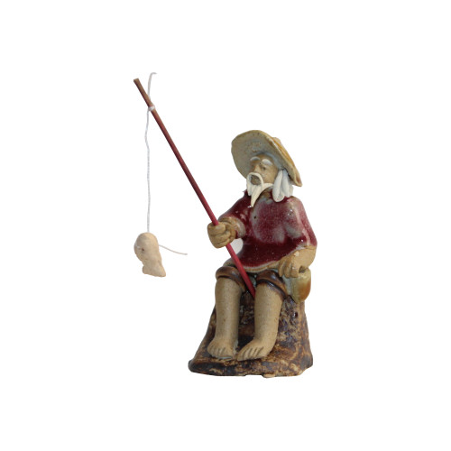 A small, detailed Chinese ceramic figurine of an elderly fisherman, commonly used as a traditional mudman decoration for bonsai trees. The figurine depicts a man with a long, flowing white beard and mustache, sitting peacefully on a textured, dark brown rocky base. He wears a wide-brimmed, conical tan hat and a tunic finished in a glossy, deep burgundy glaze, while his exposed arms, legs, and face are made of unglazed tan clay. In his hands, he holds a slender, reddish-brown fishing rod. A thin white string hangs from the tip of the rod, with a small, unglazed clay fish attached to the end, appearing as if it has just been caught. The entire piece is set against a stark white background, emphasizing its handcrafted features and tranquil theme.