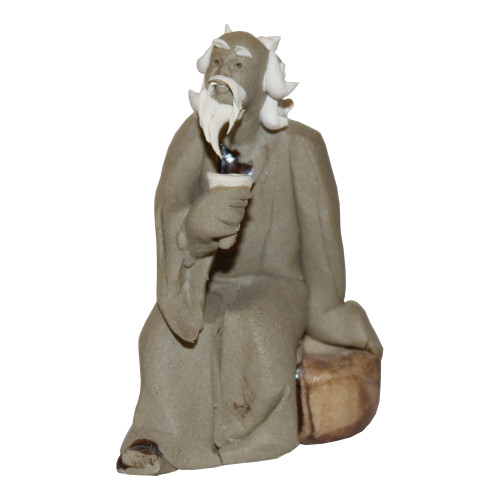 An artisanal Chinese mudman figurine (model F-090) is shown sitting on a simple, earth-toned block. The elderly figure is hunched slightly forward, holding a small, white shell-shaped cup or bowl in his right hand near his chest. He has a serene, contemplative expression with his gaze directed slightly upward. 

The figurine is crafted from traditional greyish-brown clay with a matte, textured finish that highlights the folds and wide, draped sleeves of his traditional robes. Contrasting with the earth-toned body, his long, flowing beard, mustache, and the hair on the sides of his head are finished in a smooth, bright white glaze. Fine details include dark-glazed accents under his beard and on his exposed foot. This classic bonsai accessory is often used to add a sense of scale, history, and tranquility to miniature landscapes and Zen gardens.