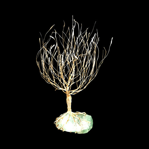An intricate, handmade silver wire tree sculpture is showcased against a stark, solid black background. The sculpture features a thick trunk made of numerous strands of gleaming silver wire twisted together, which split and fan out into a dense, rounded canopy of hundreds of fine, delicate branches reaching upwards. The base of the tree consists of silver wire roots that tightly wrap around and anchor the piece to a large, translucent, light green raw gemstone with a natural, unpolished texture. The contrast between the bright, metallic silver wire and the soft, glowing green stone creates a sophisticated and ethereal aesthetic, characteristic of high-quality bonsai-inspired wire art. This piece, labeled as NTR-08, represents a fusion of natural mineral beauty and meticulous metal craftsmanship.
