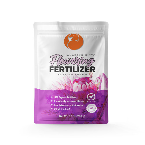 A detailed studio product shot of a 10 oz (280 g) white resealable stand-up pouch of Hanasaku Hiryo Flowering Fertilizer by Ho Yoku Products. 

The top of the bag features a distinctive orange circular logo with a stylized bird's head. Below it, the word "Flowering" is written in a large, elegant pink and white gradient cursive script, positioned above the word "FERTILIZER" in bold, black block letters. 

The lower third of the package is decorated with a vibrant purple and pink watercolor illustration of large blossoms, resembling proteas. Overlaid on this floral background are several key product features in white text: "100% Organic Fertilizer," "Dramatically increases blooms," "Slow Release over 4-6 weeks," and an "NPK of 1.5-9-4.5." 

To the right of the bullet points are two circular icons: a checkmark with the text "READY TO USE" and a "100% Satisfaction Guaranteed" seal. The bag sits against a clean, solid white background with a soft shadow underneath, emphasizing its professional packaging designed for flowering plants and bonsai maintenance.