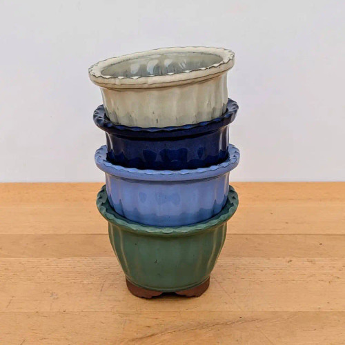 A vertical stack of four small, decorative glazed ceramic bonsai pots is arranged on a light-colored wooden tabletop against a clean white background. The collection showcases a variety of colors, featuring—from top to bottom—a cream-white, a dark navy blue, a soft periwinkle blue, and a muted sage green pot. 

Each pot is finished with a glossy glaze and distinctive design elements, including a vertically fluted body and a decorative scalloped rim. The bottom-most green pot rests on small, unglazed reddish-brown clay feet, highlighting the material and craftsmanship of these round bonsai containers. The image highlights the different color options available for this specific style of fluted ceramic pot.