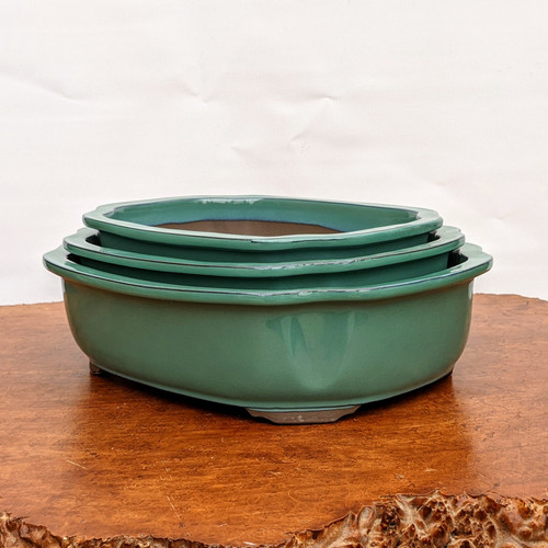 A set of three nested Oribe glazed Japanese bonsai pots from Tokoname, stacked from largest to smallest. Each pot features a rich, glossy forest green glaze with traditional variations, a characteristic of the Oribe style. The rectangular pots have elegant, rounded corners and a decorative scalloped "mokkou" (quince flower) rim. Crafted from high-quality Japanese ceramic, the pots rest on small, integrated feet and are displayed on a polished, natural live-edge wooden table against a clean, neutral white background. The image highlights the exquisite craftsmanship and deep color typical of authentic Tokoname ceramic bonsai containers.