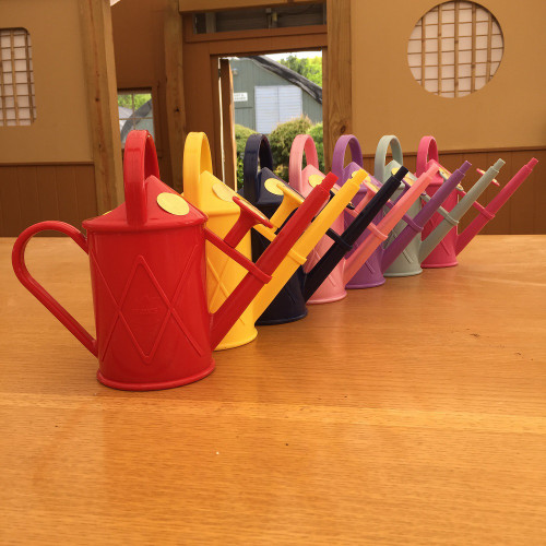 A row of seven vibrant, 2-pint plastic watering cans for bonsai, arranged diagonally on a light wood surface. The cans come in a rainbow of colors: bright red in the foreground, followed by yellow, navy blue, light pink, lavender, sage green, and hot pink. Each watering can has a classic design with a side handle, a top arch handle, and a long, slender spout. In the background, a warm, wooden interior features traditional-style elements like a circular latticed window, suggesting a peaceful garden shed or bonsai studio environment.