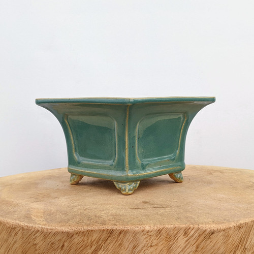 A square 8.5-inch glazed ceramic bonsai pot in a vibrant teal green color, featuring a smooth, glossy finish. The pot is designed with a wide flared rim and decorative recessed rectangular panels on each side. It rests on four small, ornate feet with intricate scrollwork details. A golden-brown glaze accentuates the edges and corners of the pot, highlighting its architectural shape. It is displayed on a thick, round wooden pedestal with a natural grain texture, set against a plain, solid white background.