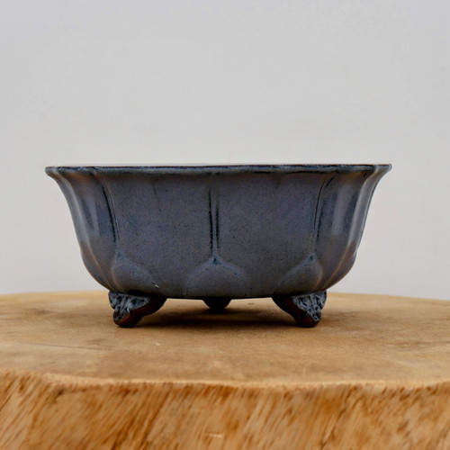A high-resolution, side-view studio photograph of a glazed Yixing bonsai pot, model YX905, resting on a thick, light-grained wooden slab. The pot is finished in a sophisticated, dusty blue-grey glaze with a subtle, fine-grained texture. Its deep, round body features elegant vertical indentations that create a lobed, petal-like effect around its circumference, leading up to a thin, slightly flared rim. The vessel is elevated on three small, ornate "cloud" feet carved from dark reddish-brown clay, which provides a beautiful contrast to the blue glaze. The minimalist, off-white background draws focus to the pot's refined craftsmanship and classic Japanese-inspired aesthetic.