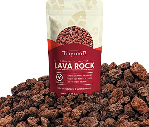 A standing resealable bag of Tinyroots Premium Soil Additive Lava Rock is centered against a white background, partially nestled in a large, scattered pile of the product. The volcanic rocks are porous, jagged, and feature natural earthy shades of reddish-brown and charcoal. 

The top half of the packaging is cream-colored with a subtle leaf pattern and a circular clear window revealing the small rocks inside. The lower half of the bag is a deep burgundy red with bold white text that reads "LAVA ROCK." Bullet points on the label highlight the product's benefits, including promoting soil drainage and plant oxygen, containing no artificial coloring, and being ideal for bonsai and succulents. The loose pile of lava rock in the foreground showcases the coarse, gritty texture of the sifted mineral, which is a key component for creating well-draining bonsai soil mixes.