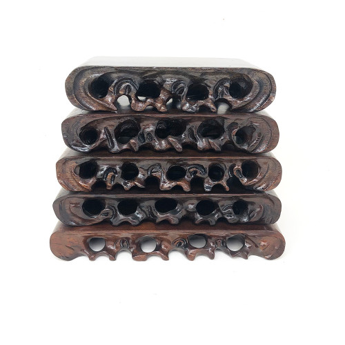 A vertical stack of five hand-carved bonsai root display stands, showcasing a range of sizes from small to large. Each stand is meticulously crafted from a rich, dark brown wood, finished with a smooth, polished sheen that catches the light. The most striking feature is the intricate, hand-carved openwork along the sides, which mimics the organic, gnarled, and winding forms of natural tree roots or weathered rock. This traditional oriental 'root carving' style adds a sense of movement and natural beauty to the pieces. The stands are stacked against a clean, white background, highlighting their deep wood tones and complex silhouettes. The top of each rectangular stand is a flat, polished surface, designed to provide an elegant and stable base for displaying a bonsai tree, a suiseki viewing stone, or other fine art objects.