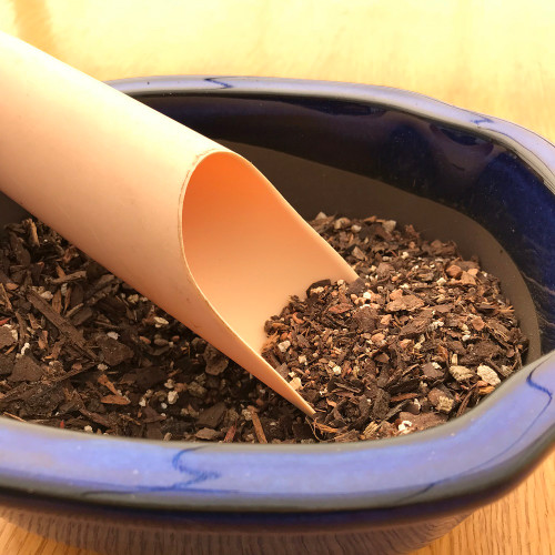 A close-up photograph featuring a light peach-colored plastic soil scoop from a 3-piece set, angled into a deep blue, glossy ceramic bonsai pot. The scoop is filled with a textured bonsai potting mix composed of dark brown bark bits and light-colored grit. The pot itself is partially filled with the same coarse soil blend. This practical demonstration shows the utility of plastic soil scoops for precise potting and repotting of bonsai trees. The scene is set on a light-colored wooden tabletop, emphasizing a clean and organized bonsai gardening workspace.