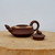 A side-profile view of a traditional, handmade Yixing teapot crafted from dark reddish-brown purple clay, or zisha. The teapot features a squat, wide body with two stylized, cream-colored floral patterns on its shoulder. The short spout has a leaf-like form, and the simple, circular C-shaped handle provides a comfortable grip. 

To the right of the main body sits the matching lid, which is uniquely adorned with a small, detailed figurine of a frog perched on top of a textured surface resembling a lotus seed pod with circular indentations. The entire set rests on a rustic, light-toned wooden cross-section against a plain, neutral white background, highlighting the artisanal craftsmanship of this functional piece of tea-ceremony art.