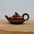A detailed side-profile view of a handmade Yixing teapot, crafted from traditional dark reddish-brown Zisha clay. The teapot features a low, wide body decorated with delicate orange-yellow lotus leaf motifs on its shoulder. A small, intricately sculpted ceramic frog sits atop the lid, serving as a whimsical handle, surrounded by small decorative perforations. The teapot's spout is uniquely designed to resemble a budding leaf, while the large, thick circular handle provides a sturdy, rustic contrast to the fine details. The vessel sits centered on a natural, light-colored wooden cross-section, emphasizing its organic and handcrafted nature, set against a clean, neutral white background.
