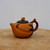 A detailed side-profile shot of a traditional Yixing clay teapot, exhibiting a warm, matte orange-brown hue. The teapot features a unique, organic shape reminiscent of a fruit or a bird, with a spout that flows seamlessly and gracefully from its body. The lid is a contrasting matte charcoal color, topped with a small, bud-like knob in the same orange-brown clay as the main body.

The handle is crafted from a darker reddish-brown clay and is textured to resemble a natural, gnarled branch, adding to the artisanal and rustic feel of the piece. Subtle decorative elements enhance its design, including a small, raised reddish-brown bat-like motif near the spout and dark, textured leaf-like appliques that curve around the body near the handle. The teapot rests on a rustic, light-colored wooden surface with a visible grain, set against a plain, neutral-toned background, which highlights its fine craftsmanship, earthy texture, and elegant form.